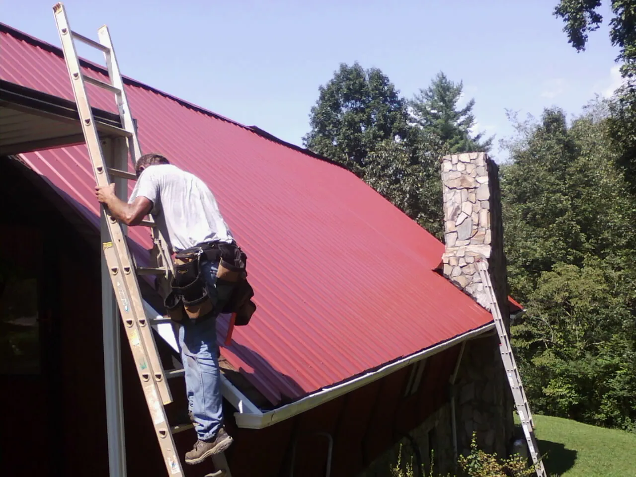 Roofer on a ladder beside a red metal roof and stone chimney — Soffit & Fascia Repair by Roofing Brothers in Fort Morgan